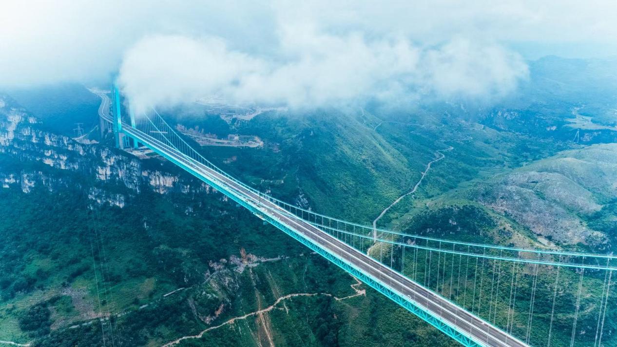 A new high-altitude suspension bridge with advanced technology, including a智慧缆索吊装系统 and多普勒激光雷达测风系统, spans across a lush mountainous valley, celebrating its opening in Guizhou's Huajiang Canyon.